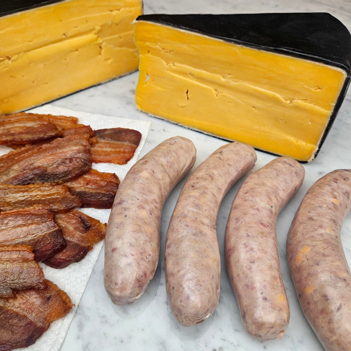 Raw grass-fed beef bacon cheeseburger sausage links on white marble with crispy cooked bacon and a wedge of aged cheddar — Wishbone Heritage Farms, Saint George, SC