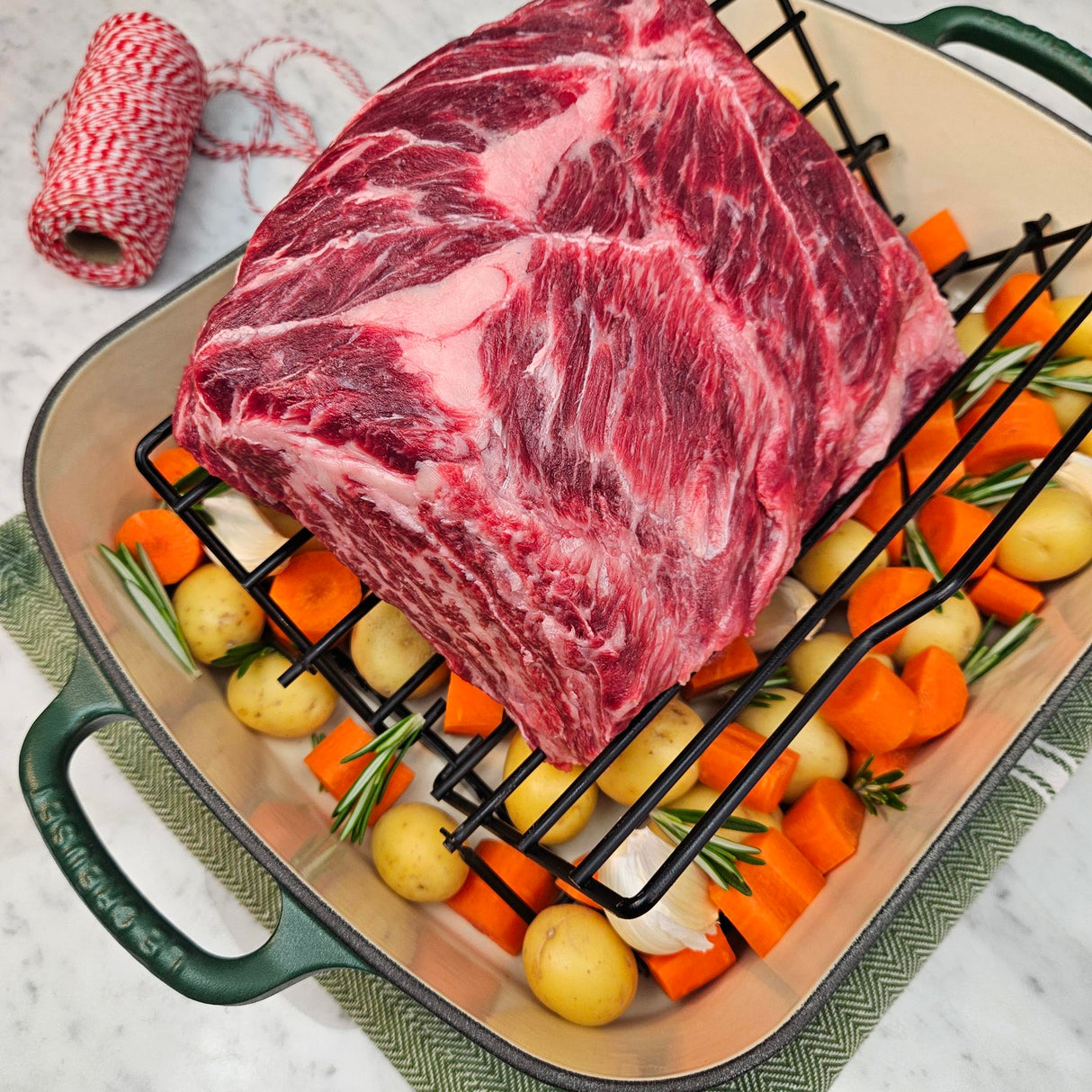 Grass-fed beef chuck eye roast in a green Le Creuset roasting pan with potatoes, carrots, garlic, rosemary, and butcher's twine — Wishbone Heritage Farms, Saint George, SC