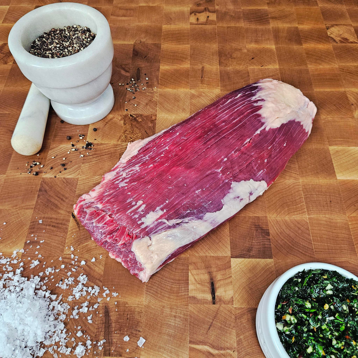 Grass-fed beef flank steak on an end-grain butcher block with flake salt, cracked pepper, and chimichurri — Wishbone Heritage Farms, Saint George, SC