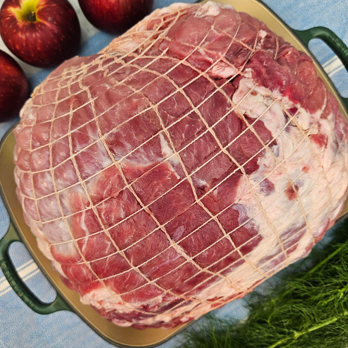 Raw pasture-raised heritage breed pork whole fresh ham, boneless and netted, filling a green roasting pan with apples and fresh fennel fronds — Wishbone Heritage Farms, Saint George, SC