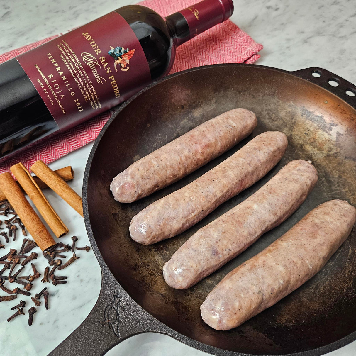 Raw pasture-raised heritage pork butifarra sausage links in a cast iron skillet with cinnamon sticks, cloves, and a bottle of Rioja — Wishbone Heritage Farms, Saint George, SC