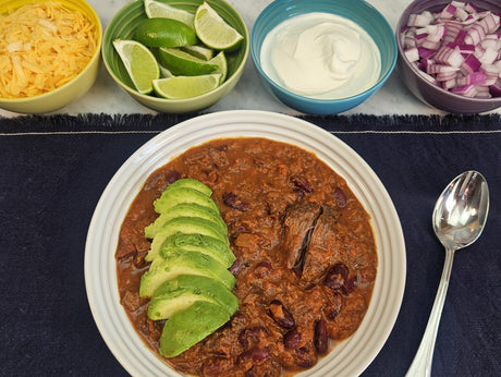 Braised beef chili topped with sliced avocado served in a white bowl on a navy linen, with toppings of shredded cheddar, lime wedges, sour cream, and diced red onion — Wishbone Heritage Farms, Saint George, SC