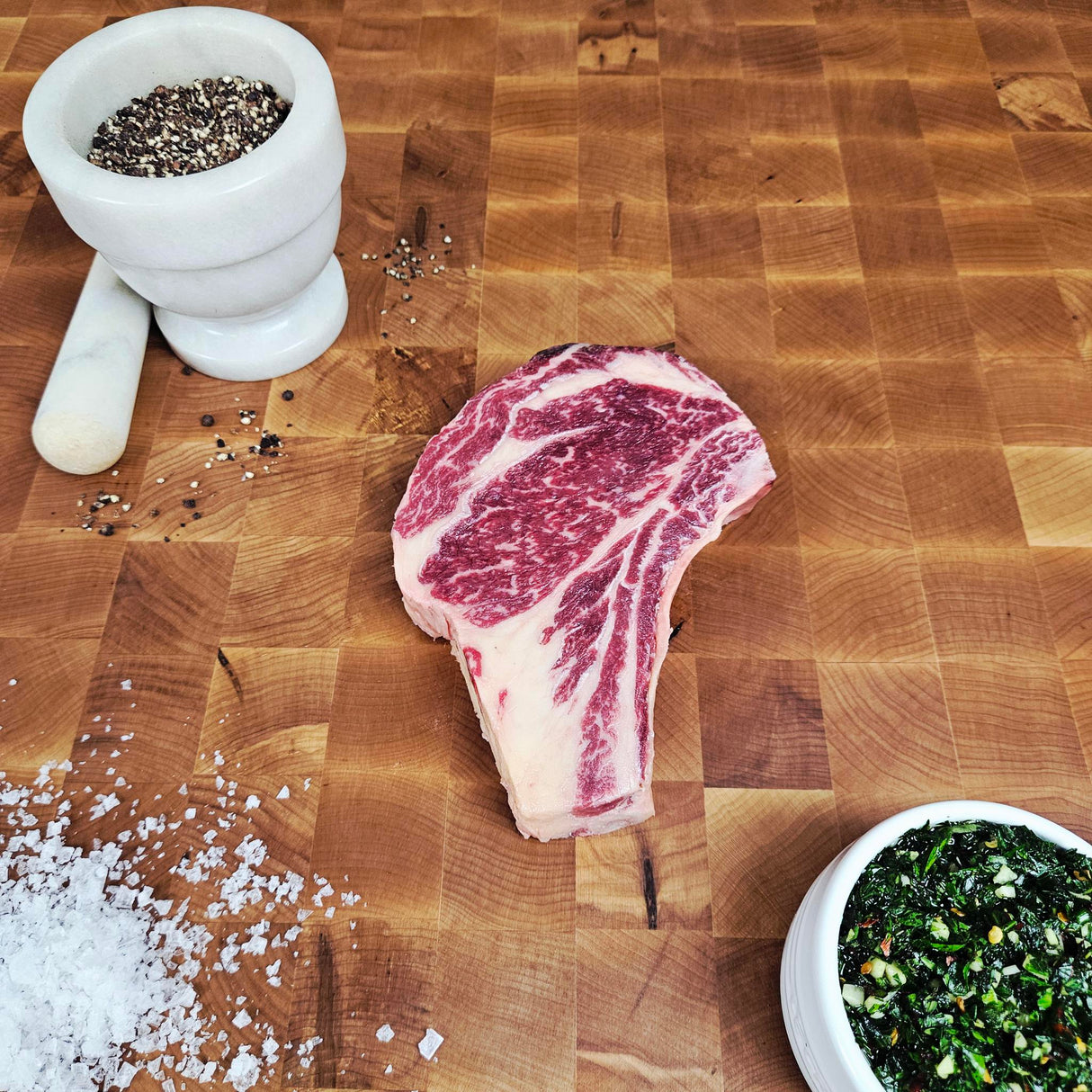 Grass-fed beef bone-in ribeye steak on an end-grain butcher block with flake salt, cracked pepper, and chimichurri — Wishbone Heritage Farms, Saint George, SC