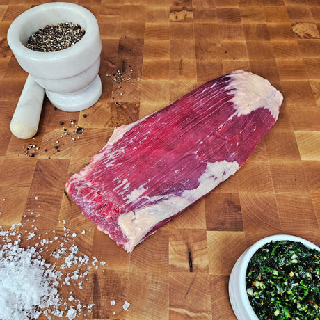 Grass-fed beef flank steak on an end-grain butcher block with flake salt, cracked pepper, and chimichurri — Wishbone Heritage Farms, Saint George, SC