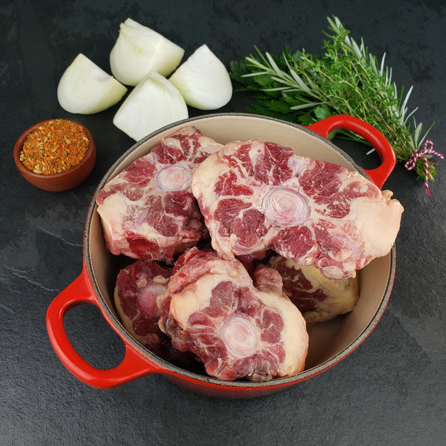 Raw grass fed beef oxtail in red Dutch oven with quartered onion, fresh herbs, and spices