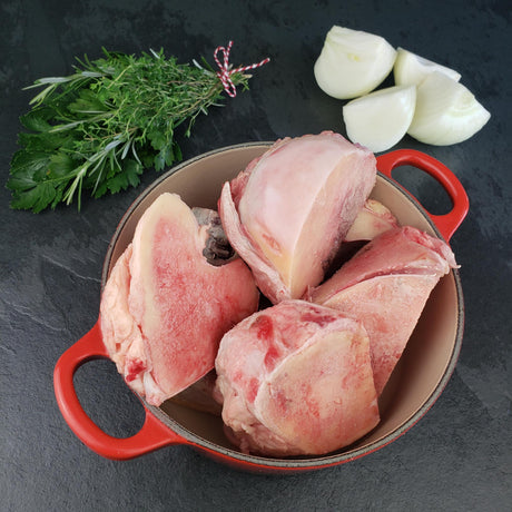 Raw grass fed beef soup bones in red Dutch oven with fresh rosemary, thyme, parsley, and quartered onion