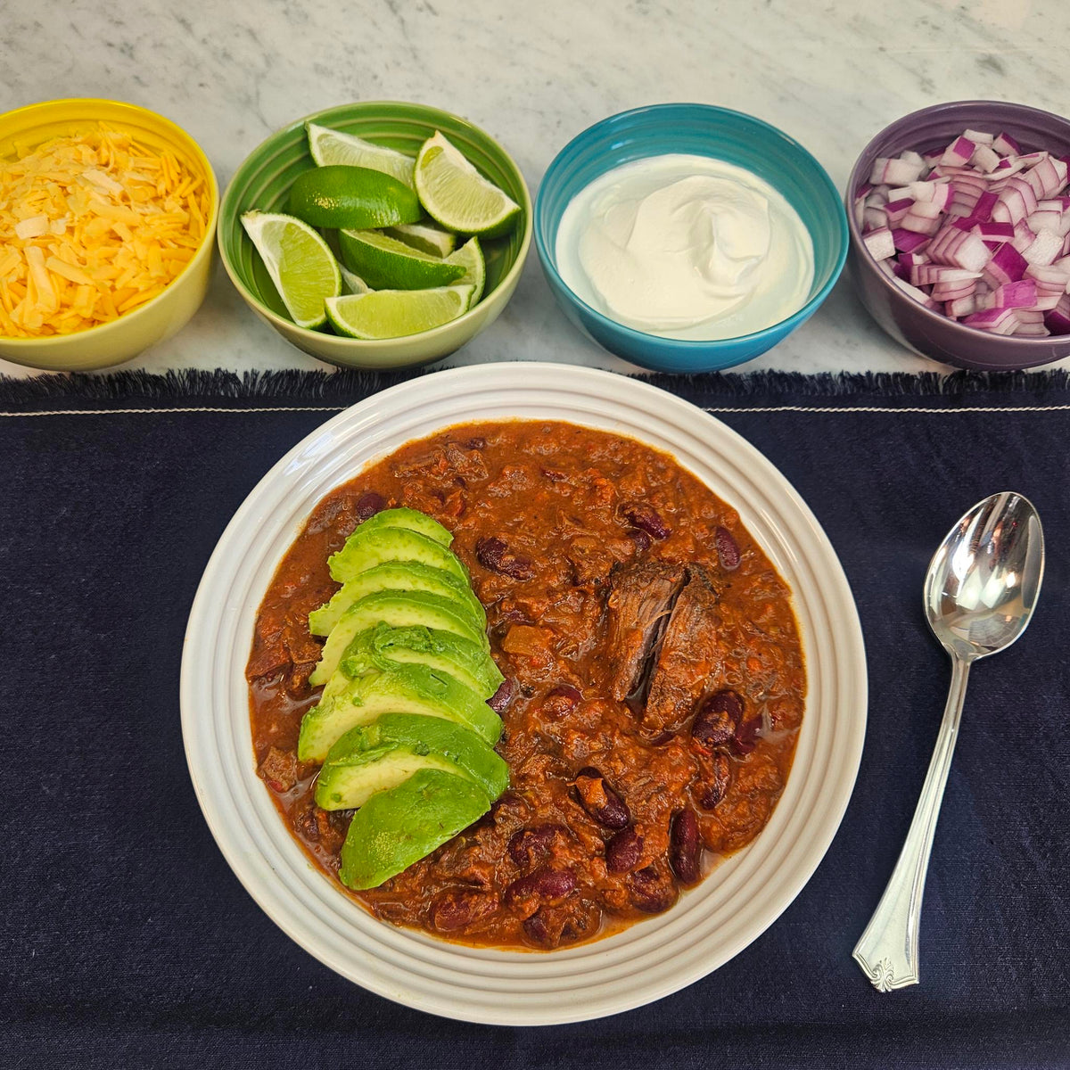 Braised beef chili topped with sliced avocado served in a white bowl on a navy linen, with toppings of shredded cheddar, lime wedges, sour cream, and diced red onion — Wishbone Heritage Farms, Saint George, SC