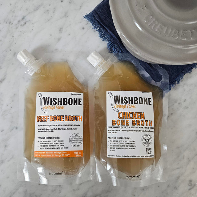 Pasture-raised chicken and grass-fed beef bone broth in 14 oz pouches on white marble with a Le Creuset saucepan — Wishbone Heritage Farms, Saint George, SC