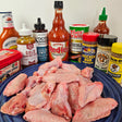 Raw pasture-raised split chicken wings on a blue plate surrounded by wing sauces and seasonings including Frank's RedHot, Sweet Baby Ray's, Lillie's Gold BBQ, Sticky Fingers Carolina Sweet, Tony Chachere's Creole, Famous Dave's Chicken Rub, Kosmos Q Wing Dust, Dan-O's Lemon Pepper, sriracha, and gochujang — Wishbone Heritage Farms, Saint George, SC