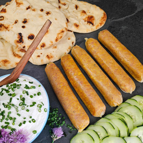 "Raw pasture-raised curry chicken sausage links on dark slate with naan, cucumber, and a chive yogurt dipping sauce — Wishbone Heritage Farms, Saint George, SC