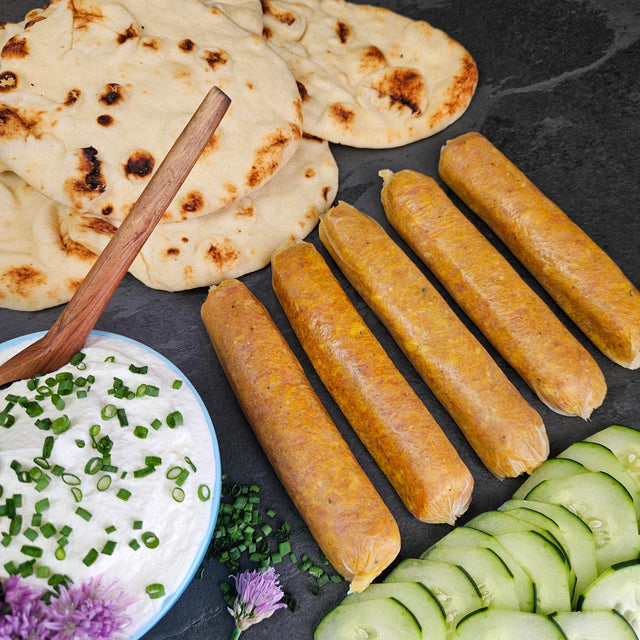"Raw pasture-raised curry chicken sausage links on dark slate with naan, cucumber, and a chive yogurt dipping sauce — Wishbone Heritage Farms, Saint George, SC