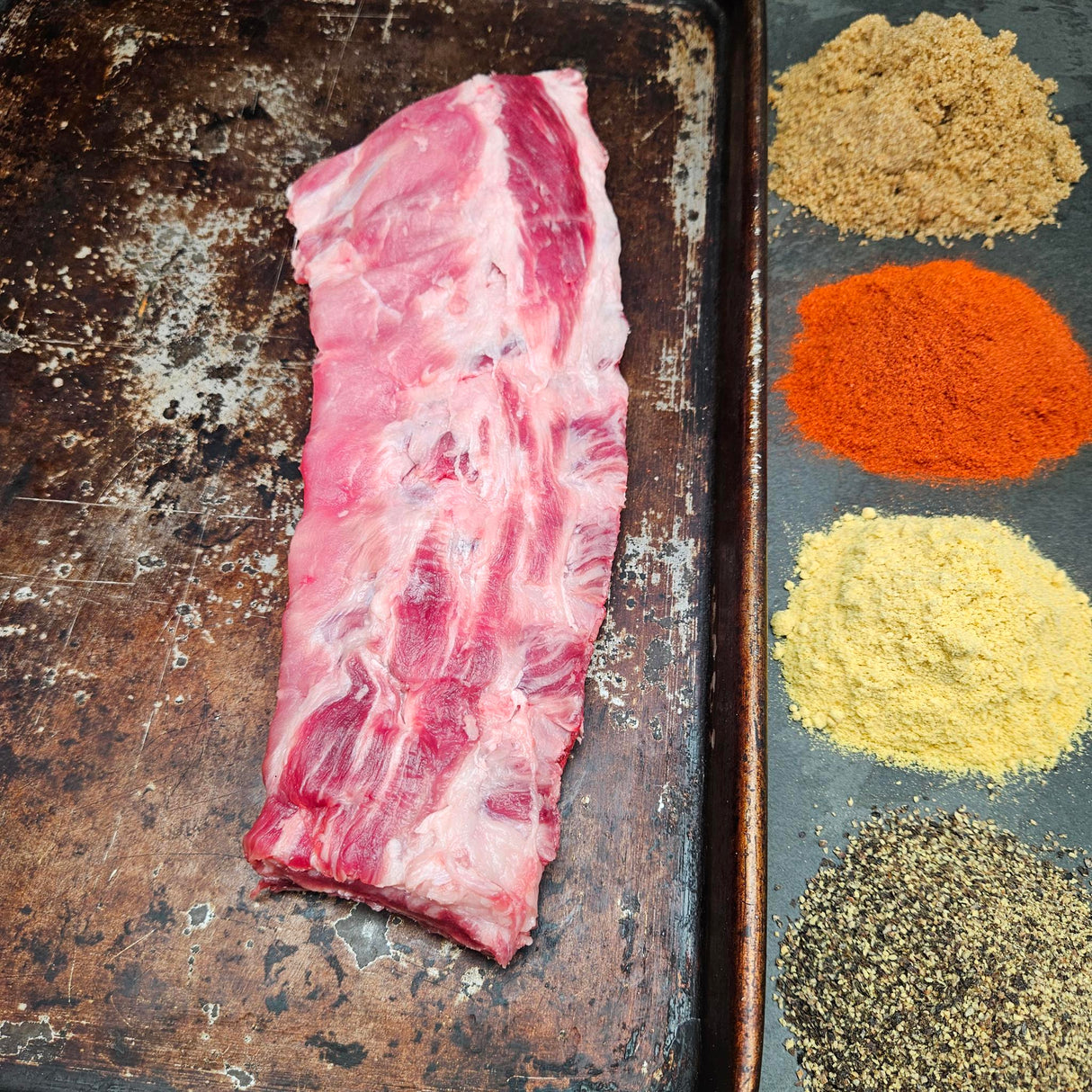 Pasture-raised heritage breed pork baby back ribs on a seasoned sheet pan with dry rub spices — brown sugar, paprika, mustard, and black pepper — Wishbone Heritage Farms, Saint George, SC