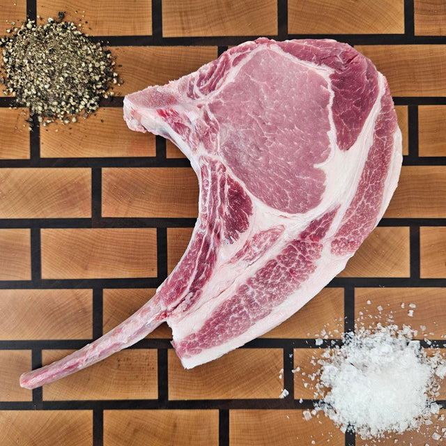 Pasture-raised heritage breed thick-cut tomahawk pork chop on an end-grain butcher block with flake salt and cracked pepper — Wishbone Heritage Farms, Saint George, SC