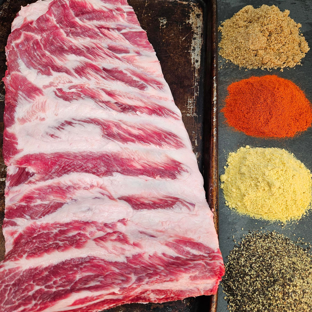 Pasture-raised heritage breed pork St Louis ribs on a seasoned sheet pan with dry rub spices — brown sugar, paprika, mustard, and black pepper — Wishbone Heritage Farms, Saint George, SC